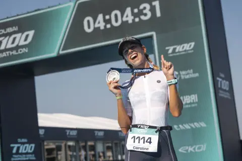 Caroline celebrates her finish at the Dubai T100 Triathlon, medal in hand, under the finish arch.