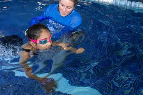 A swim instructor guides a young girl learning to swim. The question of how long does it take to learn how to swim varies, but with practice, she'll be swimming confidently.