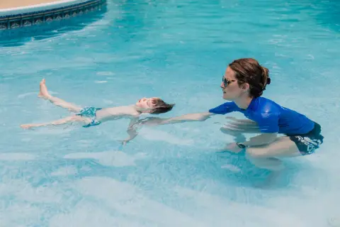 A child floats on their back in a pool during ISR swim lessons, with an instructor reaching out to support them.