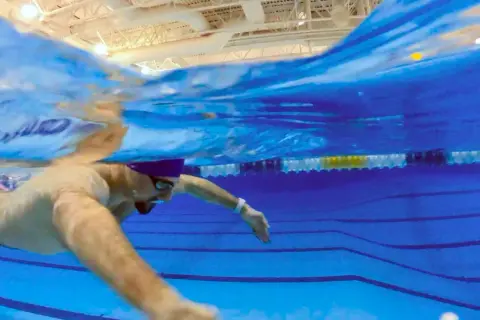 Underwater view of a swimmer in colorful trunks, demonstrating what is sculling with their arms to propel themselves through the blue pool water.