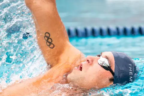 Swimmer in backstroke position, with Olympic rings tattoo on arm, mid-stroke, water splashing.