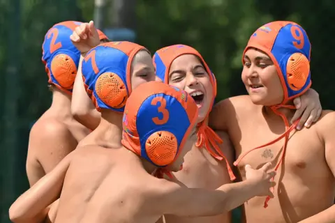 A group of young water polo players, wearing orange and blue caps with numbers, celebrate a victory. One boy, with a big smile, shouts "Habawaba!