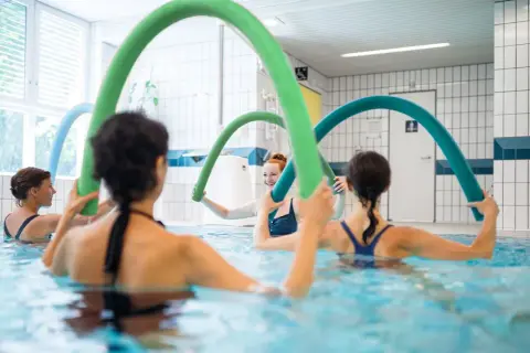 Women enjoy an aqua zumba class, holding colorful pool noodles for resistance and fun in the water.
