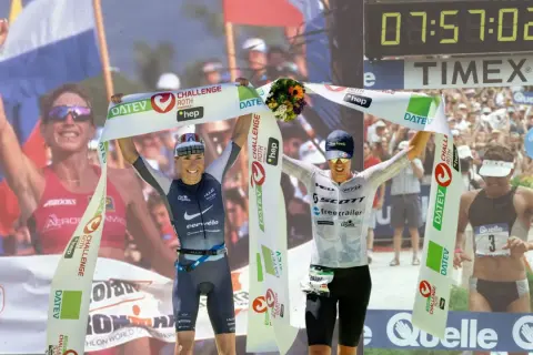 Two athletes celebrate crossing the finish line, one holding a bouquet, achieving their fastest Ironman time.