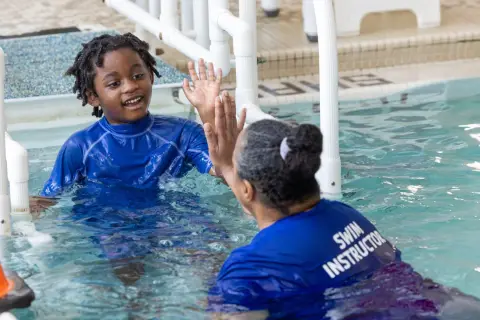 A young boy with dreadlocks in a blue rash guard gives a high-five to a swim instructor. This is a moment of connection during an autism swim lesson.