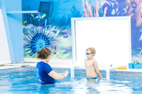 A swim instructor guides a young boy in goggles during 6 month swim lessons. The pool has a fun underwater mural.