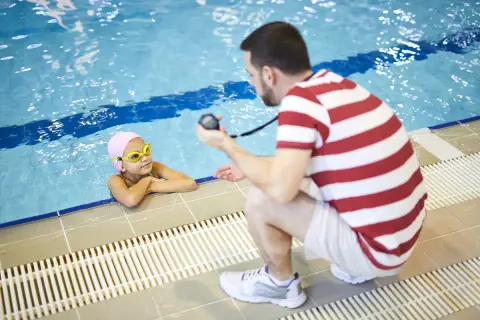 A mobile swim instructor crouches by the pool, holding a stopwatch, as a young girl in a pink cap and goggles rests her arms on the edge.