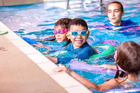 Children enjoy swim lesson games, splashing and laughing in the pool with goggles and flotation devices.