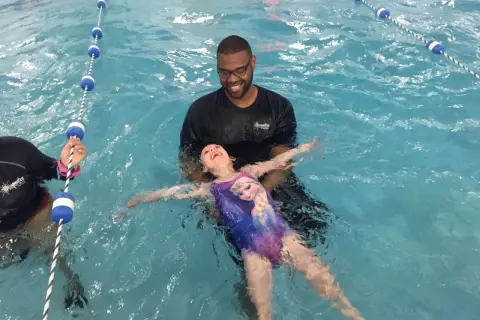 A smiling instructor helps a young girl in a Frozen swimsuit learn how to float on her back in a swimming pool.