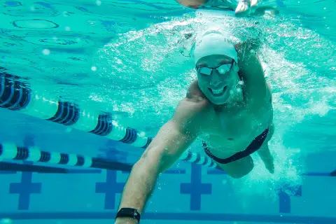 A smiling man swims underwater in a pool, enjoying easy swim workouts. Lane lines and pool markings are visible.