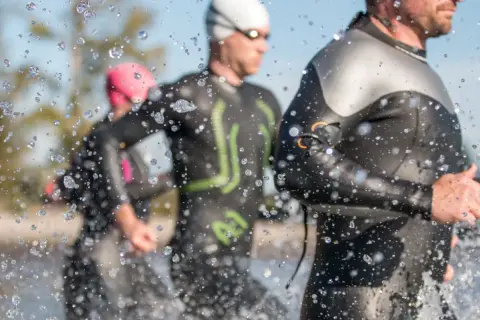 Triathletes in Ironman wetsuits run through water, splashes obscuring the view. Following Ironman wetsuit rules, they prepare for the race.