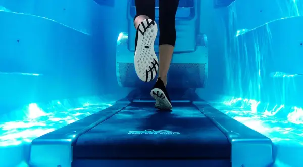 Underwater view of a person's legs and feet on a treadmill, performing aqua jogging in a bright blue pool.