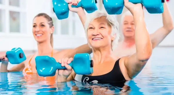 Seniors and a younger woman enjoy pool exercises, lifting blue dumbbells for a refreshing workout.