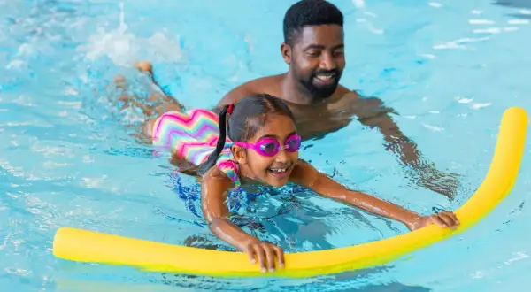 A father smiles as he helps his daughter with her swim lessons. She's wearing goggles and holding a yellow pool noodle.