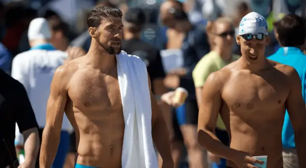 Two muscular men, one with a towel draped over his shoulder, showcase the lean physique of a swimmer body vs. a gym body.