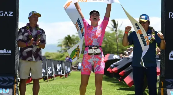 Triumphant athlete crosses the finish line at Hawaii 70.3, holding the banner high. Two men in leis congratulate her.