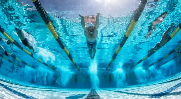 Underwater view of swimmers in a pool, with a focus on a swimmer performing butterfly stroke. The sun shines through the water, illuminating the lanes and the powerful stretches for swimmers.