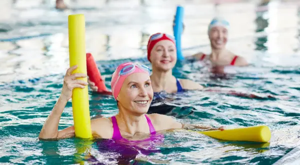 Three smiling fitness swimmers in a pool, holding yellow pool noodles.