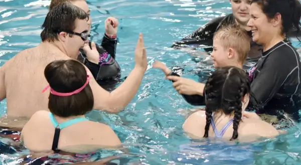 A group enjoys swim lessons for special needs, with instructors and participants high-fiving and smiling in the pool.