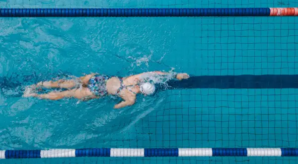 A lone swimmer glides through a pool lane, their back and arms creating splashes. The colorful swimsuit stands out against the blue water.