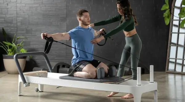 A man on a reformer uses resistance bands for pilates for swimmers, with a trainer guiding his form.