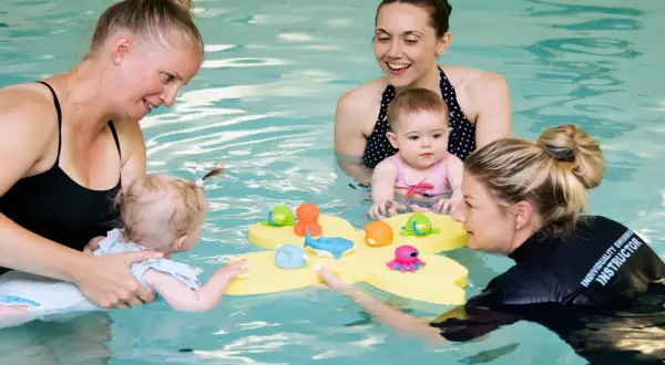Moms and instructors in a pool with toddlers, playing with bath toys. The question of how many swim lessons does a toddler need is a common one for parents.