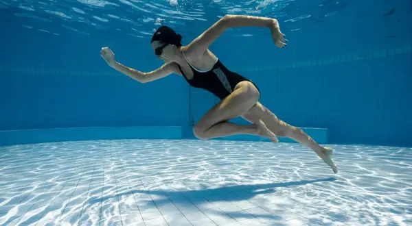 Underwater shot of a woman in a black swimsuit performing aqua aerobics. Her body is streamlined as she propels herself through the blue water.