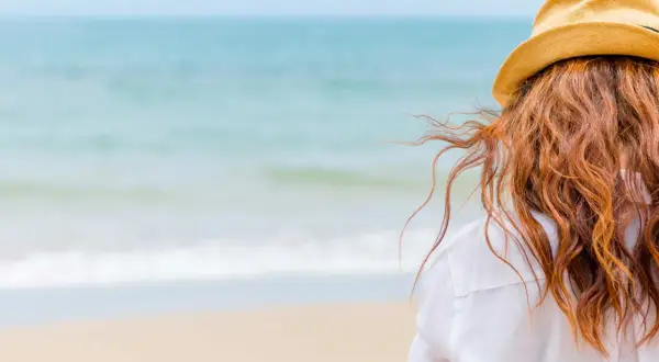 Woman with red hair in a straw hat on the beach, wondering how soon can I swim after coloring my hair.