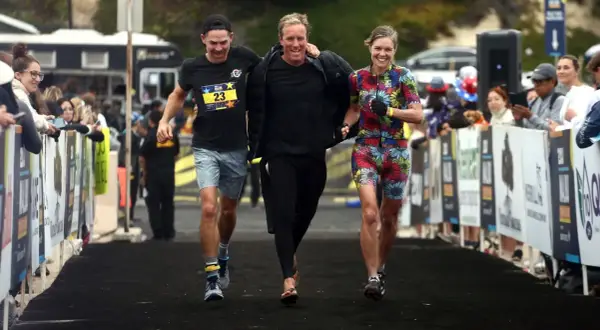 Three friends celebrate crossing the finish line of an Ironman relay. The woman in the colorful floral suit beams, while the two men, one in a black shirt with a race bib, share a laugh.