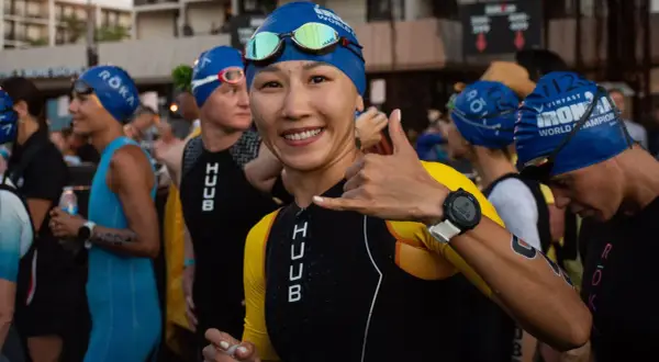 A smiling woman in a blue swim cap and yellow and black triathlon suit gives a shaka sign, ready for the honu triathlon.