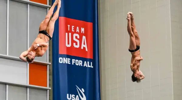 Two divers perform a synchronized high dive, suspended mid-air in a dramatic pose against a "Team USA" banner.