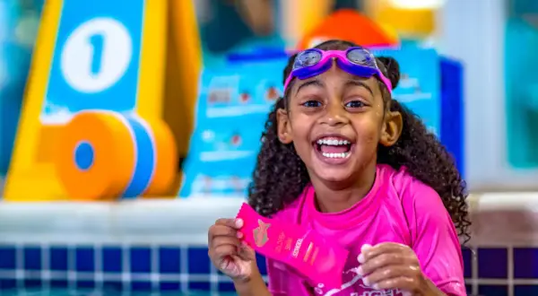 A happy girl in goggles and a pink rash guard holds a Goldfish Swim School ribbon. She's in a pool, ready for swim classes for 5 year olds.