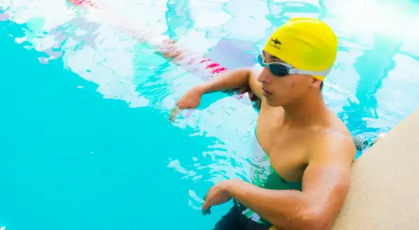 A young swimmer in a yellow cap and goggles rests by the pool edge, contemplating how much do swim instructors make.