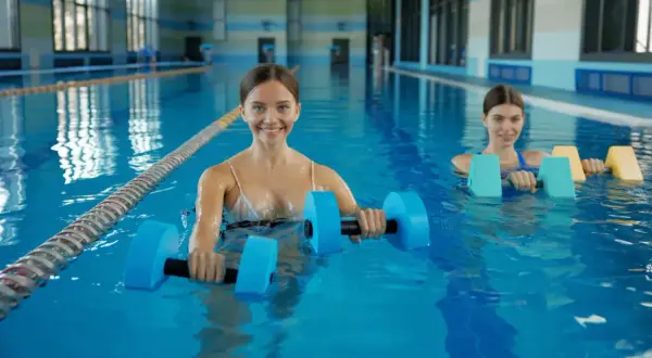 Two women enjoy a refreshing swim workout in a bright indoor pool, using buoyant dumbbells for resistance training.