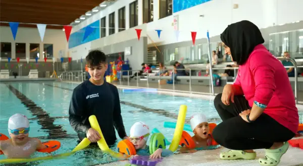 A swim instructor in a pink shirt and hijab coaches young children in a pool, demonstrating the rewarding nature of swim instructor jobs.