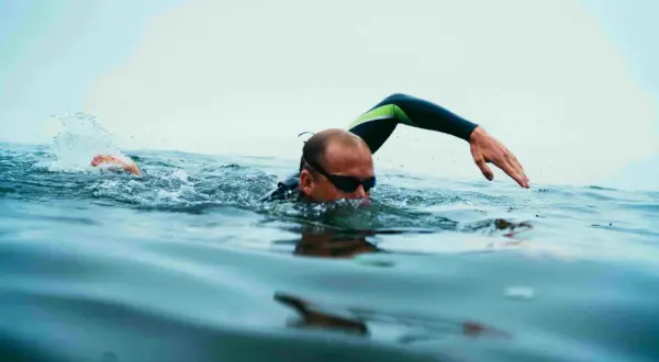 Swimmer in goggles and wetsuit performs swim drills, with one arm extended and splashing water.