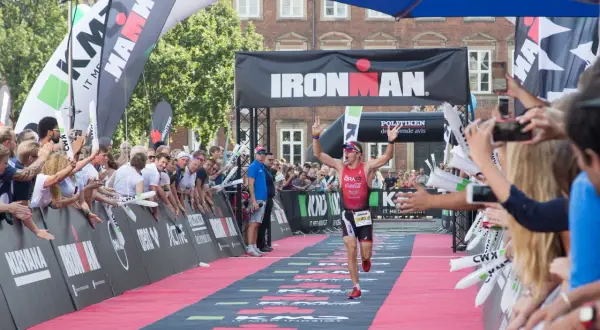 A triumphant athlete crosses the finish line at an Ironman triathlon, arms raised in victory, cheered by a crowd.
