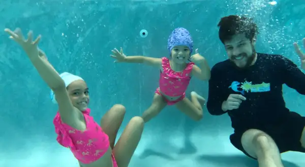 A swim instructor smiles while teaching two young girls how to teach toddler to hold breath underwater.