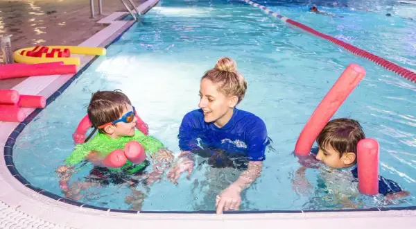 A smiling swim instructor in a blue shirt teaches two young boys in a pool. They are using pink pool noodles for support.