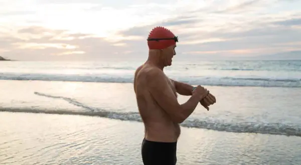 Man in swim cap checks watch on beach, ready for his swim. Early morning or late evening might be the best time to swim for weight loss.