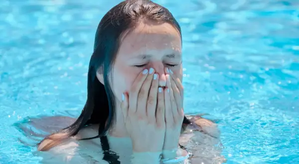 A young woman in a pool covers her nose and mouth, eyes closed, possibly experiencing a chlorine allergy sneezing fit.