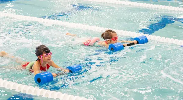 Two girls with goggles swim in a pool, holding blue flotation devices. One girl's face is partially submerged, showing her determination.