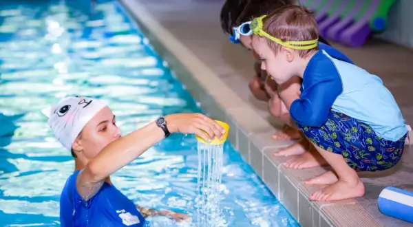 Instructor demonstrates water pouring during swim lessons for teens. Children watch intently from the poolside.