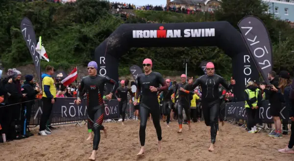 Athletes in wetsuits run from the water under an "Ironman Swim" arch. The start of their race is a sandy beach, ready for the iron man swim challenge.
