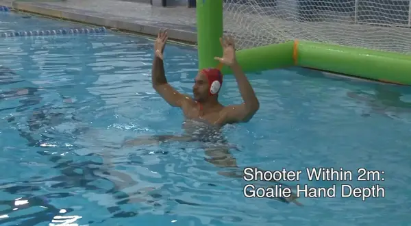 A water polo goalie in a red cap with hands raised, ready to block a shot.