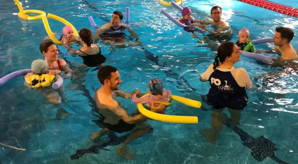 Parents and babies enjoy mommy and me swim classes, splashing and playing with pool noodles in the bright blue water.