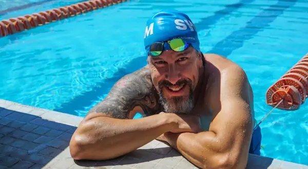 A smiling man in a blue swim cap and goggles rests on the pool edge, ready to teach how to swim laps for beginners.