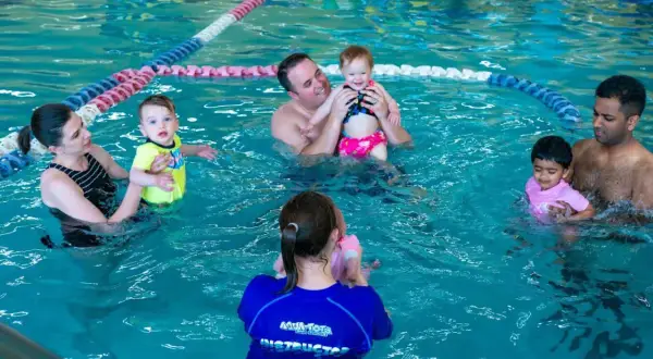 Parents and babies enjoy a swim tots class in a bright blue pool. An instructor in a blue shirt with "Aqua-Tots" visible helps a baby.