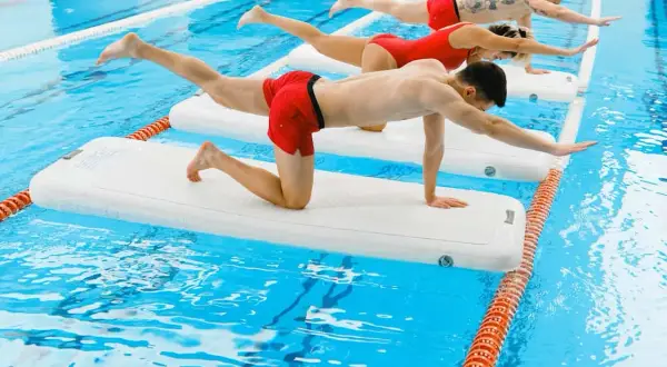 Three people practice aqua gym on inflatable mats in a pool, balancing in a plank-like pose.