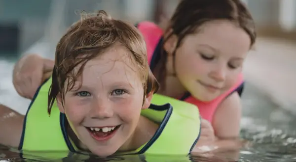 A happy boy with a missing tooth grins during 3 year old swim lessons, with a girl in a pink life vest behind him.